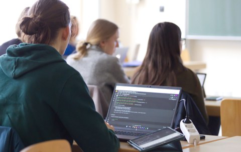 Mehrere Personen sind von hinten in einem Seminarraum zu sehen. Der Blick des Betrachter richtet sich auf einen aufgeklappten Laptopbildschirm.
