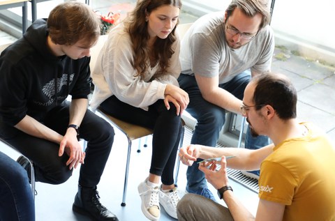 Auf dem Bild sieht man eine Gruppe aus vier Personen. Drei Personen sitzen auf Stühlen und hören einer Person zu, der ein Exponat in der Hand hält und etwas erklärt.