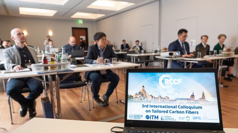 Konferenzraum mit Teilnehmenden, sitzend an Tischen und Laptop.
