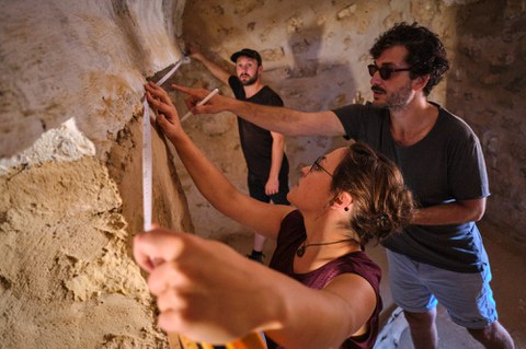 Drei Personen arbeiten zusammen, um eine raue, strukturierte Wand in einer archäologischen Stätte zu messen. Eine Person mit Brille misst die Wand mit einem Maßband, während eine andere im Hintergrund auf die Wand zeigt und die dritte Person aufmerksam zusieht.