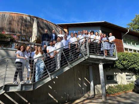 A group of people stands on an outdoor staircase in front of a building, posing for a photo. The sky is clear and blue.