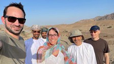 A group of six people standing together in a desert landscape with mountains in the background. They are smiling at the camera; some are wearing sunglasses, traditional clothing, and hats. The sky is clear and blue.