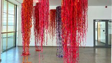 Eine Kunstinstallation mit mehreren länglichen, hängenden Strängen aus leuchtend rotem, rosa, orangefarbenem und violettem Stoff, die von der Decke in einem geräumigen Raum mit großen Fenstern und einer Glastür im Hintergrund hängen.