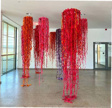 Eine Kunstinstallation mit mehreren länglichen, hängenden Strängen aus leuchtend rotem, rosa, orangefarbenem und violettem Stoff, die von der Decke in einem geräumigen Raum mit großen Fenstern und einer Glastür im Hintergrund hängen.