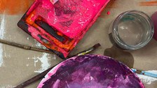Eine Draufsicht auf einen unordentlichen Malerarbeitsplatz. Es gibt einen rechteckigen Farbbehälter mit leuchtend rosa Farbe, ein Glasgefäß und eine runde Palette mit gemischter lila Farbe. Mehrere benutzte Pinsel liegen verstreut herum.