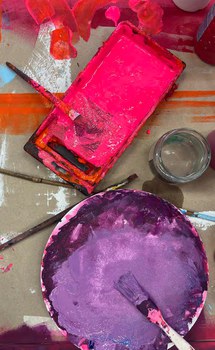 Eine Draufsicht auf einen unordentlichen Malerarbeitsplatz. Es gibt einen rechteckigen Farbbehälter mit leuchtend rosa Farbe, ein Glasgefäß und eine runde Palette mit gemischter lila Farbe. Mehrere benutzte Pinsel liegen verstreut herum.