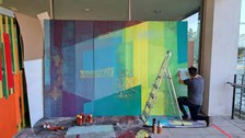 Eine Person kniet auf dem Boden und arbeitet an einem farbenfrohen Wandbild mit geometrischem Muster auf einer großen Tafel. Verschiedene Farben und Pinsel liegen verstreut herum und neben dem Wandbild steht eine Leiter.
