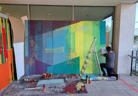 Eine Person kniet auf dem Boden und arbeitet an einem farbenfrohen Wandbild mit geometrischem Muster auf einer großen Tafel. Verschiedene Farben und Pinsel liegen verstreut herum und neben dem Wandbild steht eine Leiter.