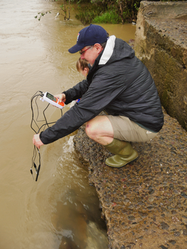 Dr. Jungmann hockt auf einem steinigen Flussufer und testet mit einem Handgerät mit angeschlossenen Sensoren das Wasser. Eine andere Person ist teilweise dahinter zu sehen, steht und beobachtet. Das Wasser erscheint trüb.