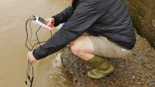 Dr. Jungmann hockt auf einem steinigen Flussufer und testet mit einem Handgerät mit angeschlossenen Sensoren das Wasser. Eine andere Person ist teilweise dahinter zu sehen, steht und beobachtet. Das Wasser erscheint trüb.