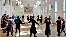 Eine Gruppe von Menschen steht in einem Kreis und hält sich an den Händen in einem gut beleuchteten Raum mit großen Fenstern und Hängelampen. Die meisten tragen schwarze Kleidung und scheinen an einem Tanz oder einer Gruppenaktivität teilzunehmen.