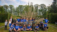 Die Gruppe der Teilnehmenden in blauen Tshirts sitzen oder stehen um ein Kunstwerk aus Bambus herum.