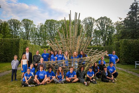 Die Gruppe der Teilnehmenden in blauen Tshirts sitzen oder stehen um ein Kunstwerk aus Bambus herum.