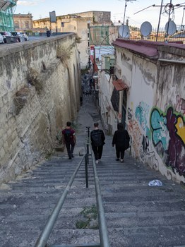 Eine schmale Steintreppe führt zwischen hohen, mit Graffiti verzierten Wänden hinab. Drei Menschen, von hinten zu sehen, gehen die Stufen hinunter. In der Ferne ist der Himmel zu sehen, und an den Gebäuden sind Satellitenschüsseln angebracht.