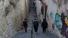 Eine schmale Steintreppe führt zwischen hohen, mit Graffiti verzierten Wänden hinab. Drei Menschen, von hinten zu sehen, gehen die Stufen hinunter. In der Ferne ist der Himmel zu sehen, und an den Gebäuden sind Satellitenschüsseln angebracht.