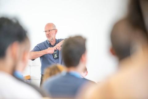 Im Vordergrund sind verschwommen die Hinterköpfe von Personen zu sehen. Scharf zu sehen ist eine Person mit Mikrofon um den Hals die gerade die Hände verschränkt.