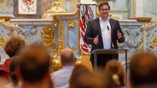 Ein Mann in Anzug und weißem Hemd spricht an einem Podium vor einem Publikum in einem großen, reich verzierten Raum. Der Hintergrund zeigt aufwendige Schnitzereien, goldene Akzente und ein Banner mit abstrakten Mustern. Im Vordergrund sind die Zuschauer.