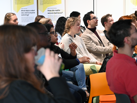 Viele Personen sitzen in Reihen im Saal und richten ihre Aufmerksamkeit nach vorne zur Bühne
