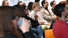 Viele Personen sitzen in Reihen im Saal und richten ihre Aufmerksamkeit nach vorne zur Bühne
