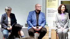 Drei Personen sitzen vorne im Raum und diskutieren moderiert ein Thema vor Publikum
