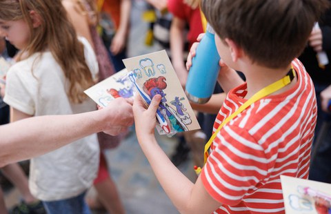 Das Foto zeigt ein Kind, was seitlich von hinten fotografiert wird und in der einen Hand eine Postkarte mit einer Figur sowie eine Zahnbürste hält. In der anderen Hand hält das Kind eine blaue Trinkflasche. Eine Hand reicht in die Mitte des Bildes eine weitere Postkarte hinein.
