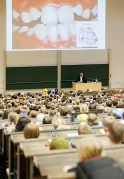 Das Foto zeigt den Teil eines Hörsaals, in dem viele Kinder sitzen. Die Perspektive der Kamera ist seitlich hinter den Kindern. Es gibt eine Referentin, die vor einer Tafel steht. Über der Tafel ist das Bild eines Gebisses zu sehen.