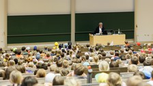 Das Foto zeigt den Teil eines Hörsaals, in dem viele Kinder sitzen. Die Perspektive der Kamera ist seitlich hinter den Kindern. Es gibt eine Referentin, die vor einer Tafel steht. Über der Tafel ist das Bild eines Gebisses zu sehen.