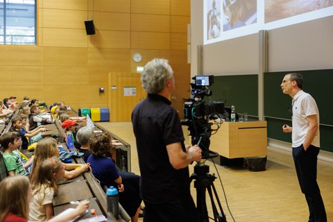 Das Foto zeigt einen Hörsaal. Auf der linken Seite sind Kinder in den Sitzreihen zu sehen. Vor den Sitzreihen hält eine männlich gelesene Person eine Vorlesung. Mittig im Foto ist ein Kameramann mit einer Kamera mit Stativ zu erkennen. 