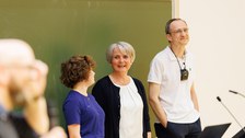 Das Foto zeigt 3 Personen, die vor einer Tafel stehen. Die Personen lachen, die Person mittig lacht in die Kamera.