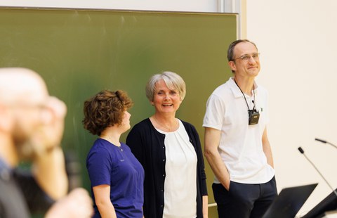 Das Foto zeigt 3 Personen, die vor einer Tafel stehen. Die Personen lachen, die Person mittig lacht in die Kamera.