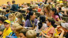 Das Foto zeigt eine Gruppe von Kindern, die sich miteinander unterhalten. Sie befinden sich in einem Hörsaal und sitzen in den Sitzreihen. Um ihnen herum befinden sich weitere Kinder.