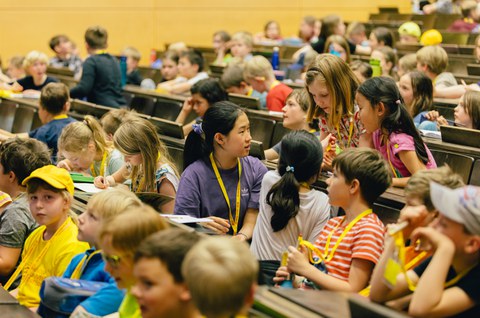 Das Foto zeigt eine Gruppe von Kindern, die sich miteinander unterhalten. Sie befinden sich in einem Hörsaal und sitzen in den Sitzreihen. Um ihnen herum befinden sich weitere Kinder.