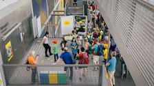 Das Foto zeigt Menschen, die vor einer Tür warten. Eine Person steht vor der Tür. Die Tür ist geschlossen. Es sind im Hintergrund Treppenstufen zu sehen.