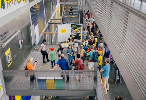 Das Foto zeigt Menschen, die vor einer Tür warten. Eine Person steht vor der Tür. Die Tür ist geschlossen. Es sind im Hintergrund Treppenstufen zu sehen.