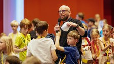 Moderator erklärt etwas, um ihn herum stehen Kinder, ein Kind meldet sich