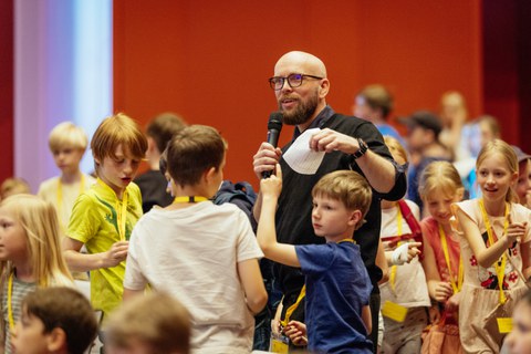 Moderator erklärt etwas, um ihn herum stehen Kinder, ein Kind meldet sich
