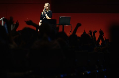 Referentin steht vor einer Menge und spricht, im Vordergrund sieht man Silhouetten von Köpfen und sich meldenden Händen