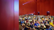 Blick in einen Vortragssaal, viele Kinder sitzen auf Stühlen und hören einem Vortrag zu