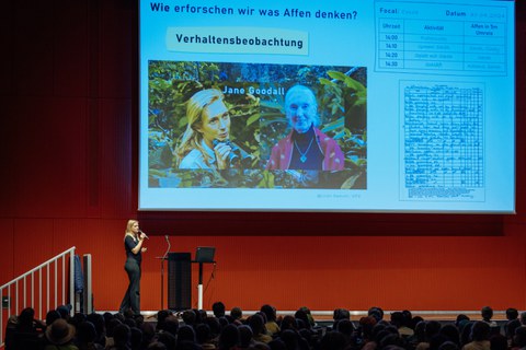 Referentin steht vor einer Menge Publikum, der Beamer zeigt eine Folie mit der Frage Wie erforschen wir, was Affen denken? und 2 Fotos von Jane Goodall sowie Aufzeichnungen einer wissenschaftlichen Studie zum Verhalten von Affen