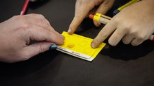 Kinder-Uni-Ausweis mit einem Stempel, 3 Hände sind zu sehen, 1 Hand hält einen Kuli