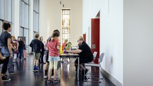 Blick auf einen Gang mit Tischen, es stehen einige Personen an den Tischen an, eine Person sitzt hinter dem vorderen Tisch