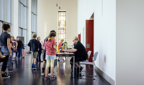 Blick auf einen Gang mit Tischen, es stehen einige Personen an den Tischen an, eine Person sitzt hinter dem vorderen Tisch