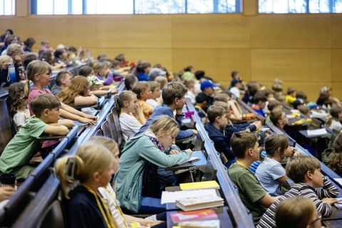 Viele Kinder sitzen in den Reihen eines Hörsaals und schauen nach vorne.