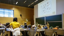 Viele Kinder sitzen in einem Hörsaal. Vor ihnen werden Folien einer Präsentation an die Wand geworfen.