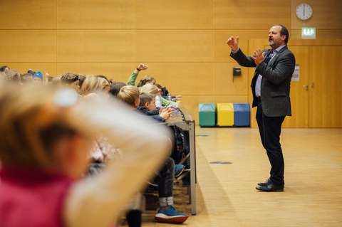 Ein Mann steht vor einem Publikum aus Kindern und erklärt etwas.