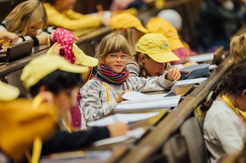 Kinder sitzen in den Sitzreihen eines Hörsaals und machen Notizen.
