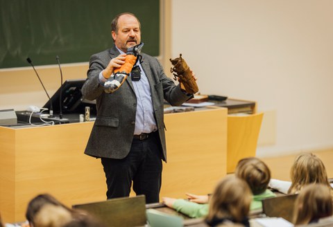 Ein Mann steht vor einer Tafel und einem Pult in einem Hörsaal und hält zwei Stofftiere in der Hand.