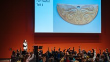 Viele Kinder sitzen im Saal. Die Referentin steht vorne und hält ihre Vorlesung. Dafür hat sie auch eine Präsentation an die Wand geworfen.