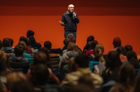 Moderator Frank Schmidt steht vor den sitzenden Kindern auf der Bühne und begrüßt sie zur Vorlesung.