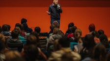 Moderator Frank Schmidt steht vor den sitzenden Kindern auf der Bühne und begrüßt sie zur Vorlesung.
